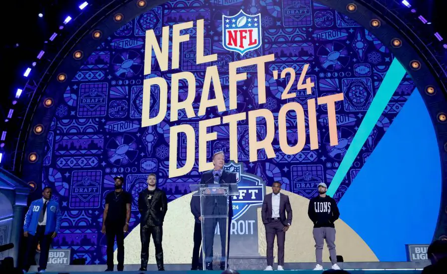 FootballR - NFL - Ein NFL Draft 2024 Event in Detroit mit einer großen, lebendigen Bühne, auf der in fetten Buchstaben „NFL Draft '24 Detroit“ steht. Ein Redner steht an einem Podium, flankiert von Figuren in Detroit Diese Beschreibung wurde automatisch generiert.